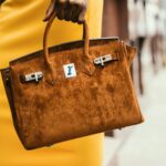 How should clothes be paired with bags? Chic brown leather handbag held by a fashionable woman in a vibrant cityscape.