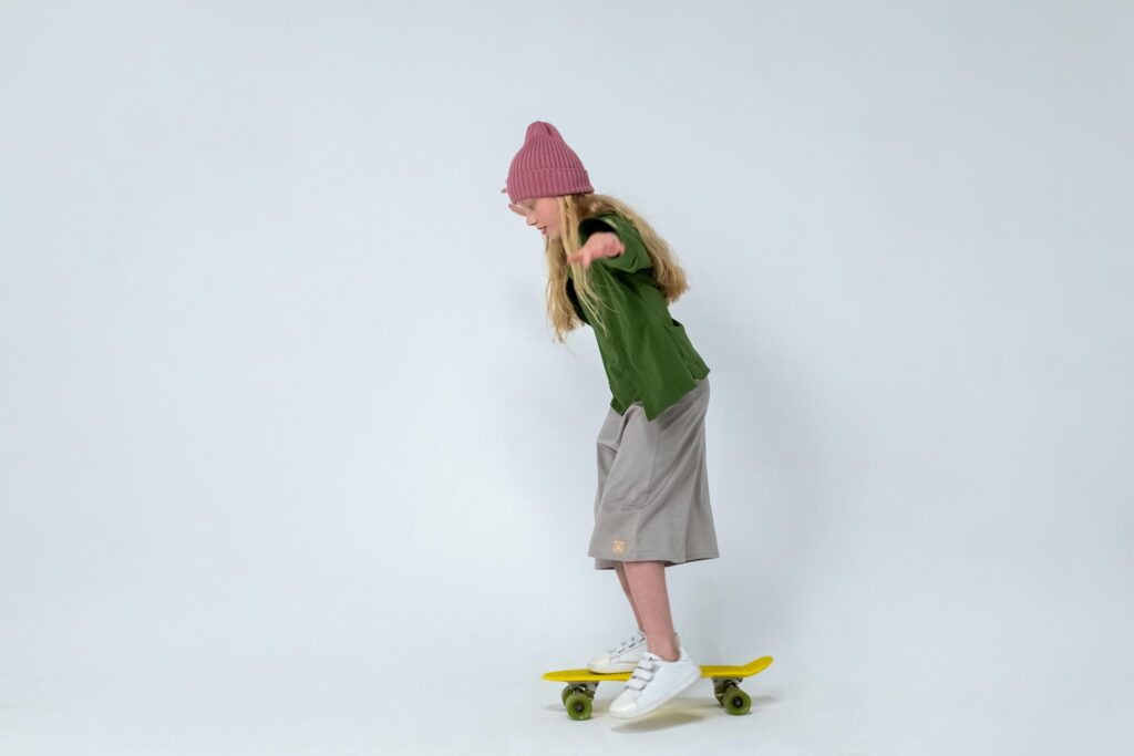 Young girl practicing skateboarding indoors, wearing a pink hat on a white background.